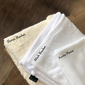 Puede incluir: Tres mantas blancas dobladas sobre una superficie de madera. La manta superior est&aacute; etiquetada como "Arctic Fleece", la manta del medio est&aacute; etiquetada como "Mink Blanket" y la manta inferior est&aacute; etiquetada como "Sherpa Blanket".