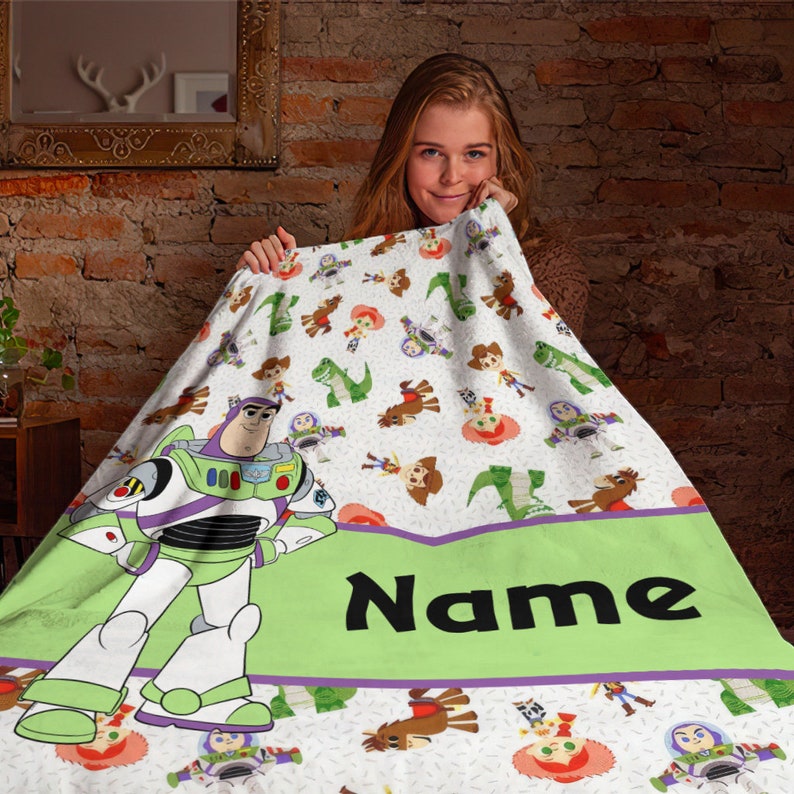 May include: A white fleece blanket featuring a repeating pattern of Toy Story characters including Buzz Lightyear, Woody, and Rex. A large Buzz Lightyear figure stands on the left, and a wide light green horizontal band displays the word Name.