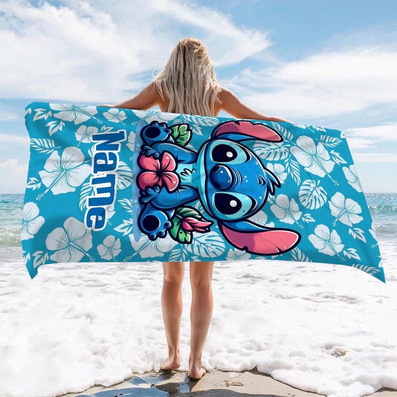 May include: A blue beach towel featuring a cartoon character with large eyes, pink ears, and a flower. The towel has a white floral pattern on a blue background. The word "Name" is printed vertically on the towel.