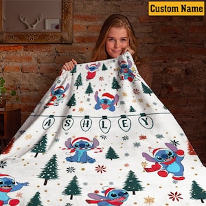 May include: A white fleece blanket featuring a repeating pattern of the blue character Stitch wearing a red Santa hat, interspersed with green pine trees, snowflakes, and a string of light bulbs spelling the name ASHLEY in black letters.