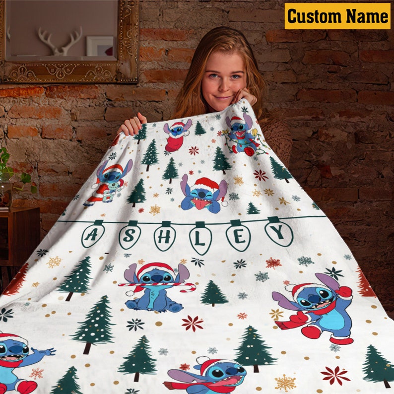May include: A white fleece blanket featuring a repeating pattern of the blue character Stitch wearing a red Santa hat, interspersed with green pine trees, snowflakes, and a string of light bulbs spelling the name ASHLEY in black letters.