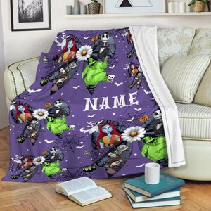 Puede incluir: Una manta morada con personajes de Pesadilla antes de Navidad. La manta tiene im&aacute;genes de Jack Skellington, Sally, Oogie Boogie y otros personajes. La palabra "NAME" est&aacute; impresa en blanco.