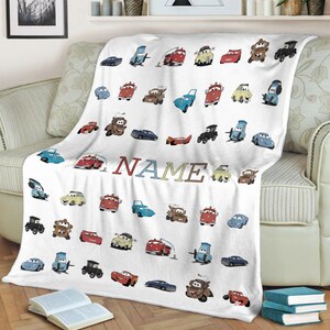 K&ouml;nnte beinhalten: Eine wei&szlig;e Fleecedecke mit einem sich wiederholenden Muster aus verschiedenen bunten Cartoon-Autos und Fahrzeugen aus dem Film Cars. Das Wort NAME ist in der Mitte in gro&szlig;en, mehrfarbigen Blockbuchstaben aufgedruckt. Die Decke hat eine weiche Textur.