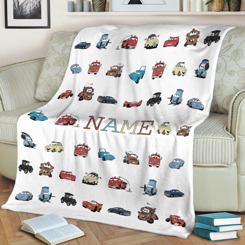 K&ouml;nnte beinhalten: Eine wei&szlig;e Fleecedecke mit einem sich wiederholenden Muster aus verschiedenen bunten Cartoon-Autos und Fahrzeugen aus dem Film Cars. Das Wort NAME ist in der Mitte in gro&szlig;en, mehrfarbigen Blockbuchstaben aufgedruckt. Die Decke hat eine weiche Textur.