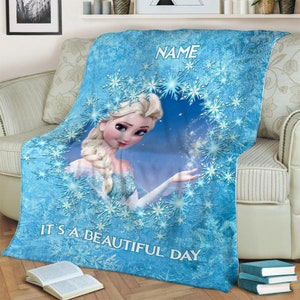 Pu&ograve; includere: Una coperta in pile blu con un'illustrazione centrale di un personaggio biondo con acconciatura a treccia, circondato da un motivo circolare di fiocchi di neve bianchi. Il testo NAME appare in alto e IT'S A BEAUTIFUL DAY in basso.