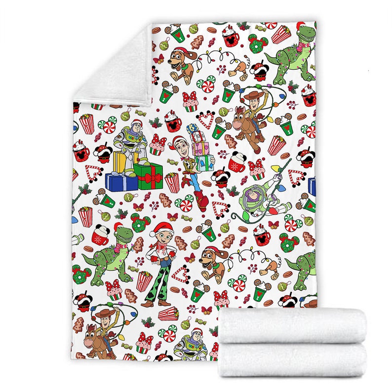 Puede incluir: Una manta de forro polar blanca con un patr&oacute;n navide&ntilde;o repetitivo de personajes de Toy Story como Woody, Buzz Lightyear, Jessie, Rex y Slinky Dog, intercalados con iconos festivos coloridos como regalos, bastones de caramelo y cacao.