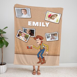 May include: A brown fleece blanket with a corkboard design featuring a cartoon image of Woody from Toy Story holding a camera. The corkboard has several photos of babies pinned to it with the name "EMILY" written in white letters.