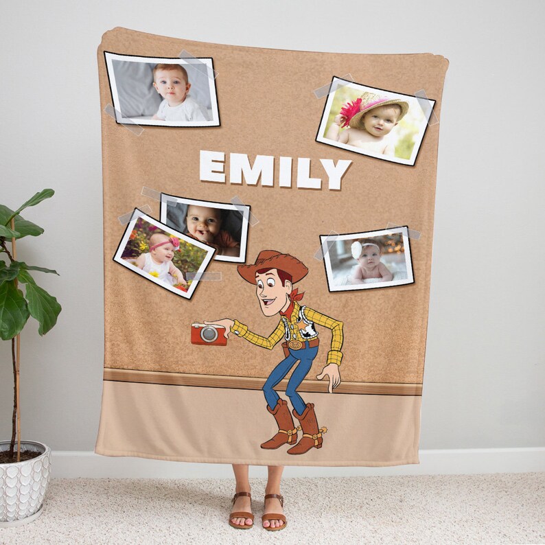 May include: A brown fleece blanket with a corkboard design featuring a cartoon image of Woody from Toy Story holding a camera. The corkboard has several photos of babies pinned to it with the name "EMILY" written in white letters.