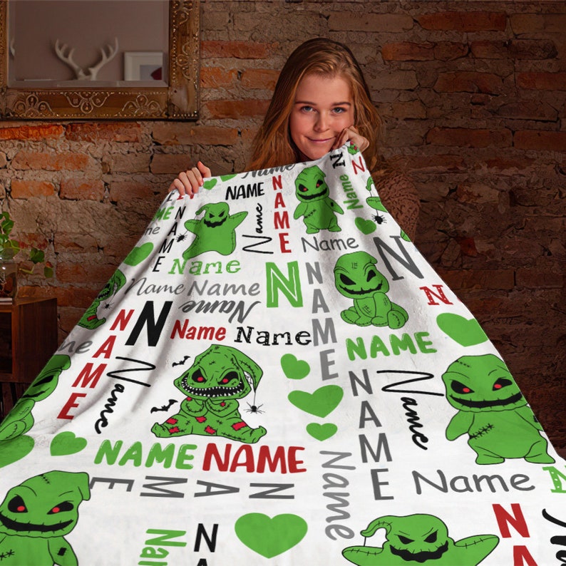 May include: A white fleece blanket with a green and red pattern featuring a cartoon character with a stitched-up face. The blanket has the word "Name" repeated throughout the design.