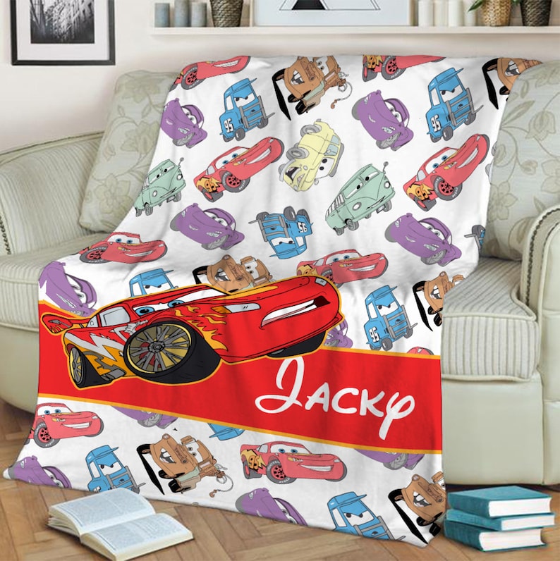 Pu&ograve; includere: Una coperta in pile bianca con un motivo ripetuto di auto dei cartoni animati in rosso, blu, viola e beige. Una grande auto da corsa rossa &egrave; raffigurata sopra una fascia rossa con la scritta bianca Jacky in corsivo.
