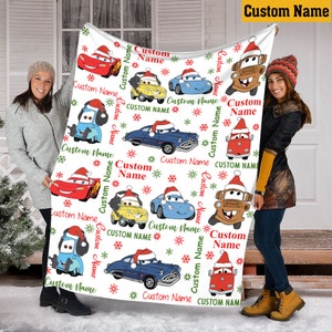 Puede incluir: Una manta de forro polar blanca con un patr&oacute;n repetitivo de coches de dibujos animados con gorros de Pap&aacute; Noel, copos de nieve rojos y verdes, y el texto Custom Name impreso en rojo y verde. La manta tiene forma rectangular y textura suave.