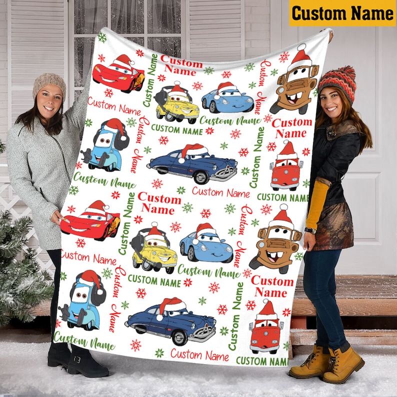 Puede incluir: Una manta de forro polar blanca con un patr&oacute;n repetitivo de coches de dibujos animados con gorros de Pap&aacute; Noel, copos de nieve rojos y verdes, y el texto Custom Name impreso en rojo y verde. La manta tiene forma rectangular y textura suave.