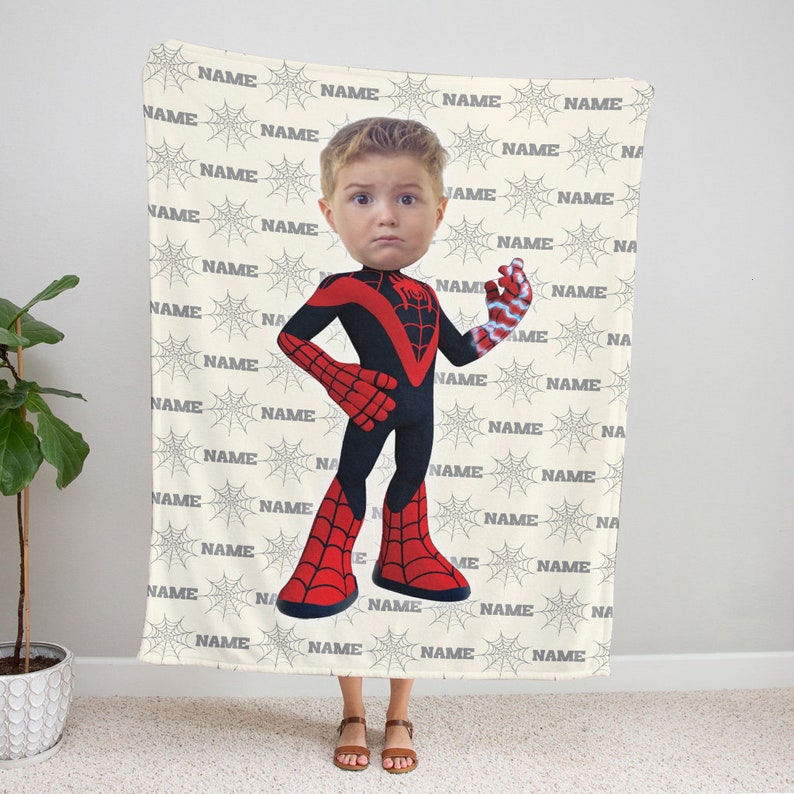 May include: A rectangular fleece blanket featuring a repeating pattern of grey spiderwebs and the word NAME in black text. Centered on the blanket is a graphic of a child's face on a superhero body wearing a black and red spider-themed suit.