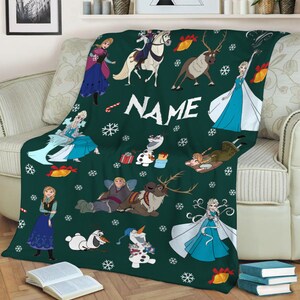 May include: A dark green throw blanket featuring a repeating pattern of Frozen characters including Elsa, Anna, Olaf, Kristoff, and Sven, interspersed with white snowflakes, red candy canes, and yellow bells, with the word NAME printed in white.