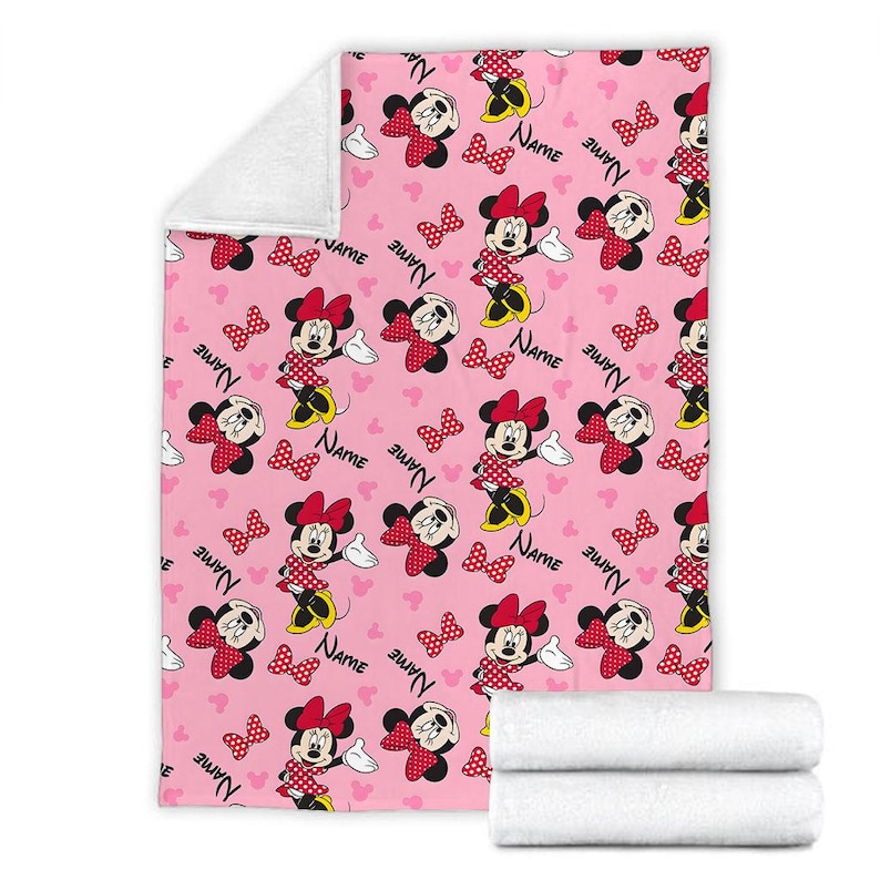 Puede incluir: Manta rosa con un patr&oacute;n repetido de Minnie Mouse, lazos rojos y la palabra "Name". La manta tiene un reverso de felpa blanca y est&aacute; doblada con dos toallas blancas.