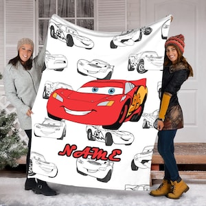 May include: A white fleece blanket featuring a large, colorful illustration of the red race car Lightning McQueen from the movie Cars, surrounded by multiple black and white line art sketches of the same character, with the red text NAME below.