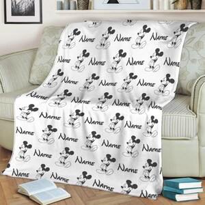 May include: A white fleece blanket featuring a repeating black and white pattern of Mickey Mouse figures and the word Name printed in a cursive script. The blanket is draped over a sofa, displaying the consistent character and text design throughout.