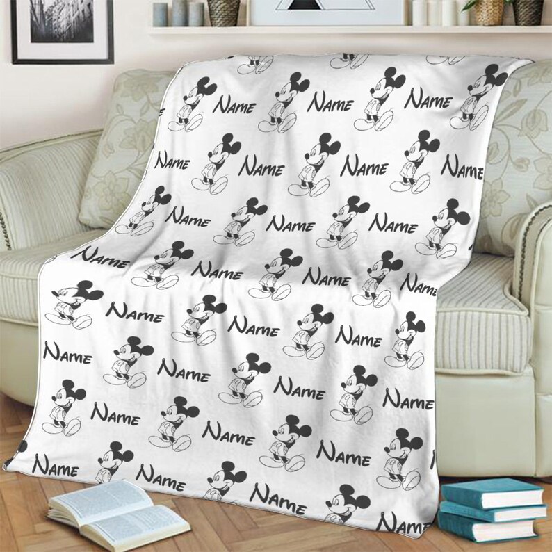 May include: A white fleece blanket featuring a repeating black and white pattern of Mickey Mouse figures and the word Name printed in a cursive script. The blanket is draped over a sofa, displaying the consistent character and text design throughout.