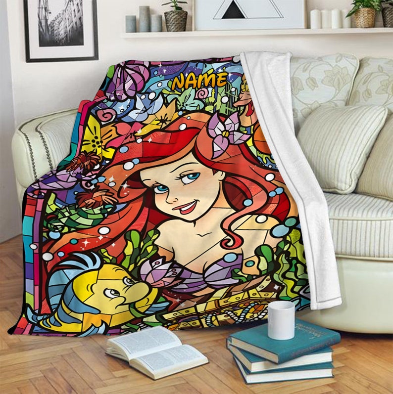 Puede incluir: Una manta de forro polar suave con una ilustraci&oacute;n colorida estilo vidriera que muestra a una sirena de pelo rojo y un pez amarillo y azul. El dise&ntilde;o incluye el texto NAME en la parte superior, rodeado de patrones acu&aacute;ticos y florales.