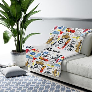 May include: A white throw blanket featuring a colourful repeating pattern of cartoon cars, tow trucks, traffic lights, steering wheels, wrenches, and road signs. The blanket includes a prominent white horizontal band with the word Name in black.