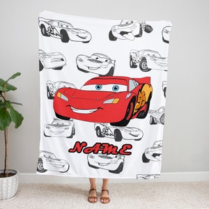 May include: A white fleece blanket featuring a large, colorful red race car with blue eyes and the number 95 on its side, surrounded by multiple black and white line art sketches of the same car in various poses, with the red text NAME below.