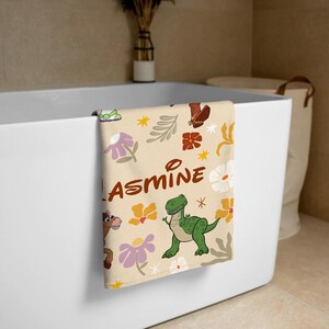 May include: A cream-colored towel featuring a repeating pattern of cartoon characters, including a green dinosaur and a brown horse, alongside stylized flowers, leaves, and stars. The name JASMINE is printed in large, brown, rounded letters across the center.