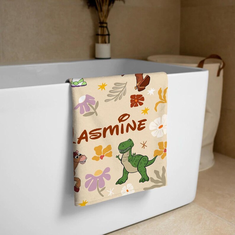 May include: A cream-colored towel featuring a repeating pattern of cartoon characters, including a green dinosaur and a brown horse, alongside stylized flowers, leaves, and stars. The name JASMINE is printed in large, brown, rounded letters across the center.