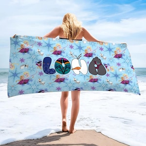 May include: A light blue beach towel featuring a pattern of snowflakes, cartoon characters, and the word "LOHA" in colorful letters. The towel is held up on a beach with the ocean and sky in the background.