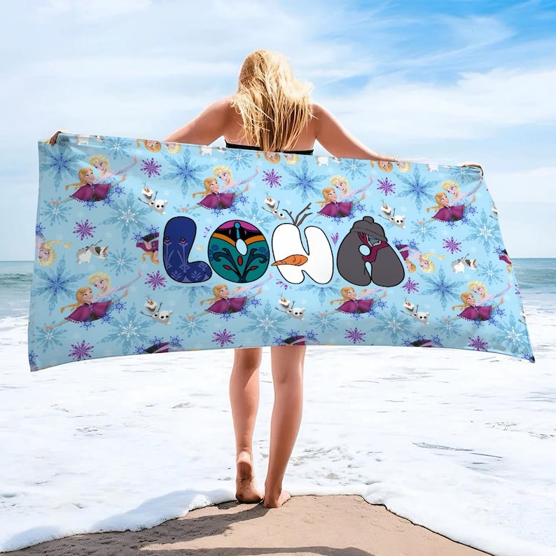 May include: A light blue beach towel featuring a pattern of snowflakes, cartoon characters, and the word "LOHA" in colorful letters. The towel is held up on a beach with the ocean and sky in the background.