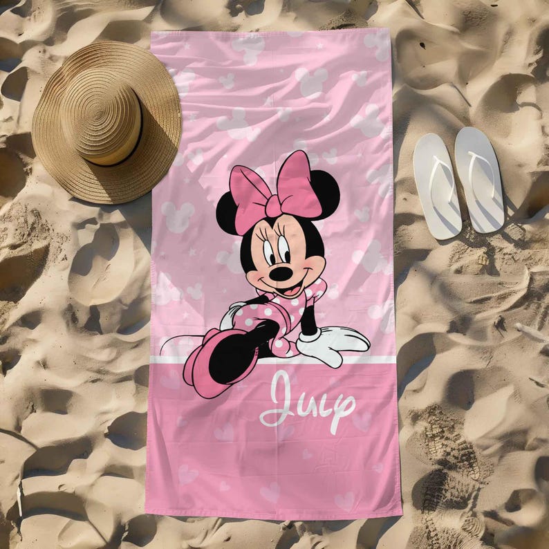 Puede incluir: Una toalla de playa rectangular con fondo rosa y siluetas blancas de Minnie Mouse, una ilustraci&oacute;n central de Minnie Mouse con vestido de lunares y lazo rosa, y el texto en cursiva blanca "July" en la parte inferior.