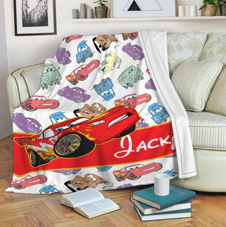 Pu&ograve; includere: Una coperta in pile bianca con un motivo ripetuto di varie auto dei cartoni animati del film Cars. Una grande auto da corsa rossa &egrave; mostrata sopra una fascia rossa orizzontale con il nome Jack scritto in corsivo bianco.