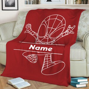 May include: A red fleece blanket featuring a white line art illustration of a superhero character in a dynamic pose. The design includes a horizontal white banner across the center with the word Name printed in a bold white sans-serif font.