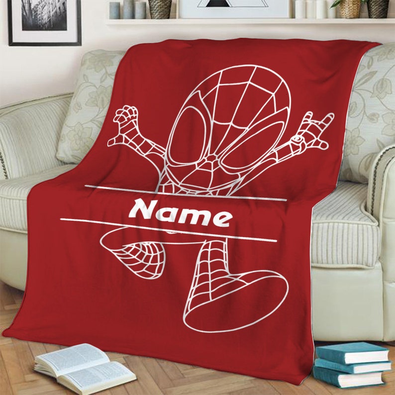 May include: A red fleece blanket featuring a white line art illustration of a superhero character in a dynamic pose. The design includes a horizontal white banner across the center with the word Name printed in a bold white sans-serif font.