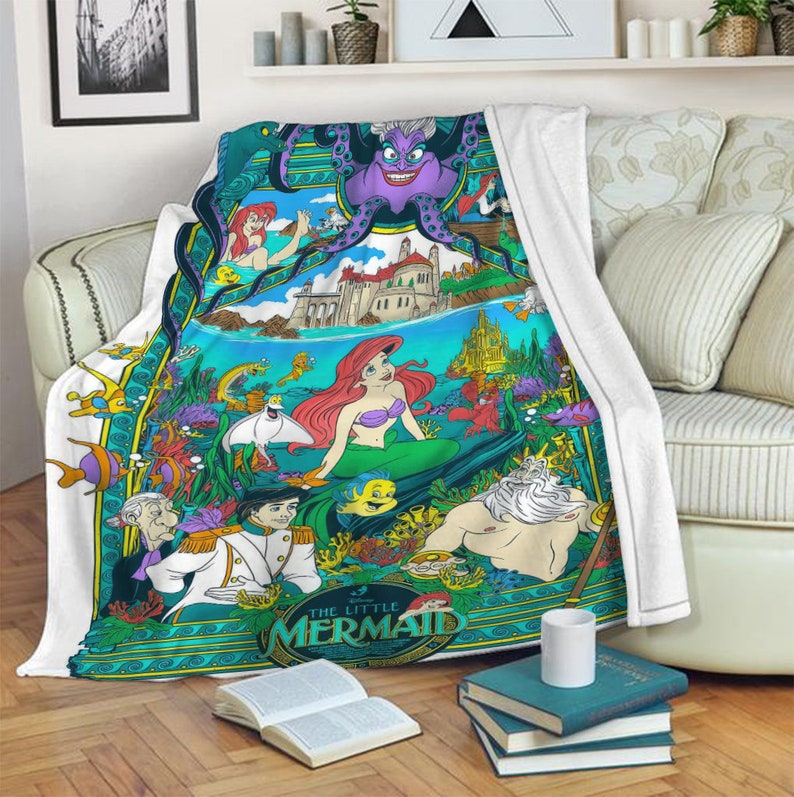 May include: A white fleece throw blanket featuring a colorful graphic print of The Little Mermaid characters including Ariel, Prince Eric, King Triton, Ursula, and Flounder. The design includes an underwater scene with a castle and coral.