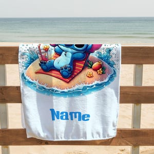 May include: White beach towel featuring a cartoon character relaxing on a tropical island. The character is blue with large ears and sunglasses, lounging on a red and orange striped towel. A drink and fruit are nearby. The word "Name" is printed in blue.