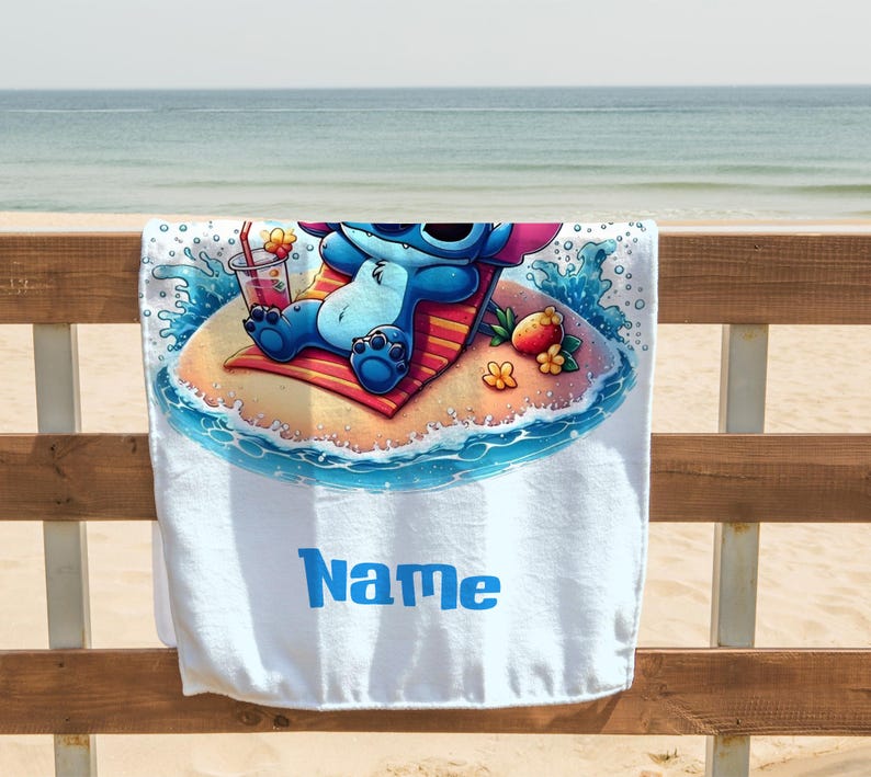 May include: White beach towel featuring a cartoon character relaxing on a tropical island. The character is blue with large ears and sunglasses, lounging on a red and orange striped towel. A drink and fruit are nearby. The word "Name" is printed in blue.