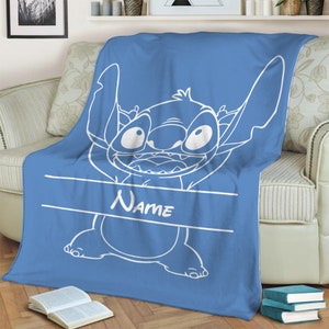 May include: A blue fleece blanket featuring a large white line art illustration of the character Stitch from Lilo and Stitch. The design includes a horizontal white banner across the center with the word Name printed in a white script font.