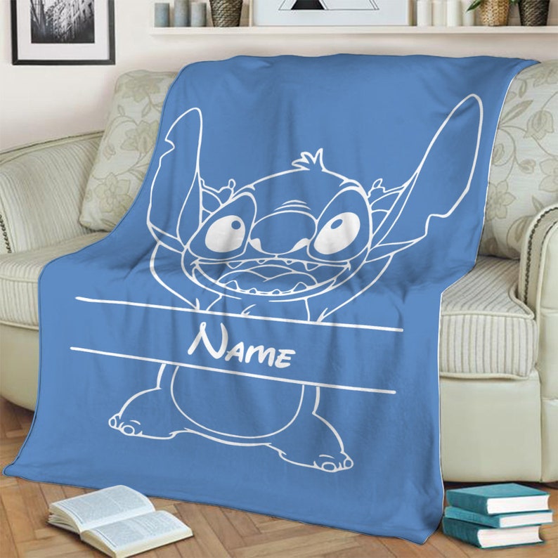 May include: A blue fleece blanket featuring a large white line art illustration of the character Stitch from Lilo and Stitch. The design includes a horizontal white banner across the center with the word Name printed in a white script font.