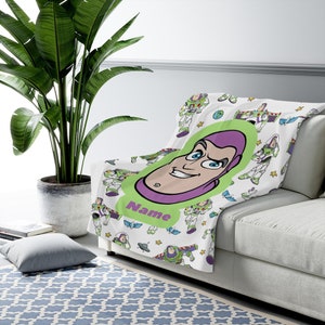May include: A white fleece throw blanket featuring a repeating pattern of Buzz Lightyear in various poses, stars, and planets. A large central green oval contains a cartoon illustration of Buzz Lightyear's face with the purple text Name below.