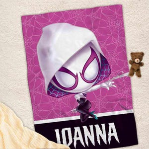 K&ouml;nnte beinhalten: Eine rosa-wei&szlig;e Fleece-Decke mit einem Cartoon-Bild einer wei&szlig;en Spinnenfrau in einem schwarzen Anzug und einem Spinnennetz-Design. Die Decke tr&auml;gt den Namen "JOANNA" in wei&szlig;en Buchstaben unten.