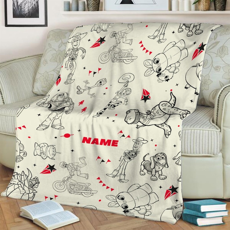 Peut inclure: Une couverture en polaire cr&egrave;me avec un motif r&eacute;p&eacute;titif en lignes noires repr&eacute;sentant des personnages de Toy Story, dont Woody, Buzz l'&Eacute;clair et Rex. Le design inclut des accents rouges en forme d'&eacute;toiles et de triangles, avec le mot NAME en rouge.