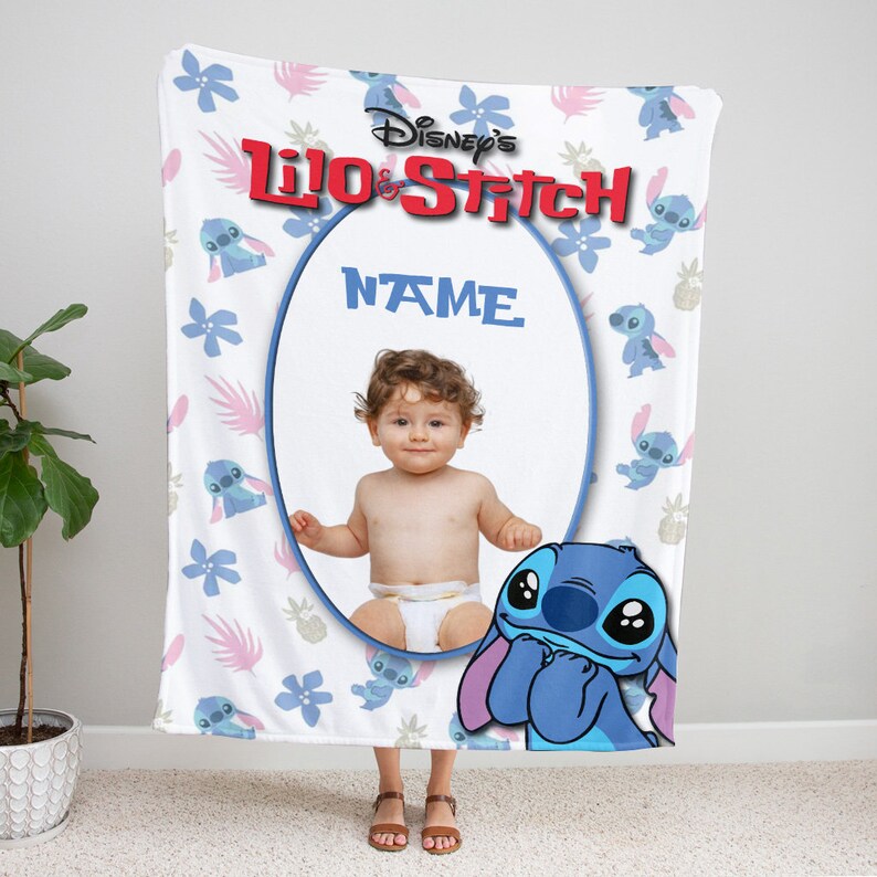 K&ouml;nnte beinhalten: Eine wei&szlig;e Fleecedecke mit einem wiederkehrenden Muster aus blauen Stitch-Figuren, rosa Palmbl&auml;ttern und blauen Blumen. Oben ist das rote Disney's Lilo & Stitch-Logo zu sehen. Ein ovaler Rahmen in der Mitte zeigt ein Babyfoto und das Wort NAME.