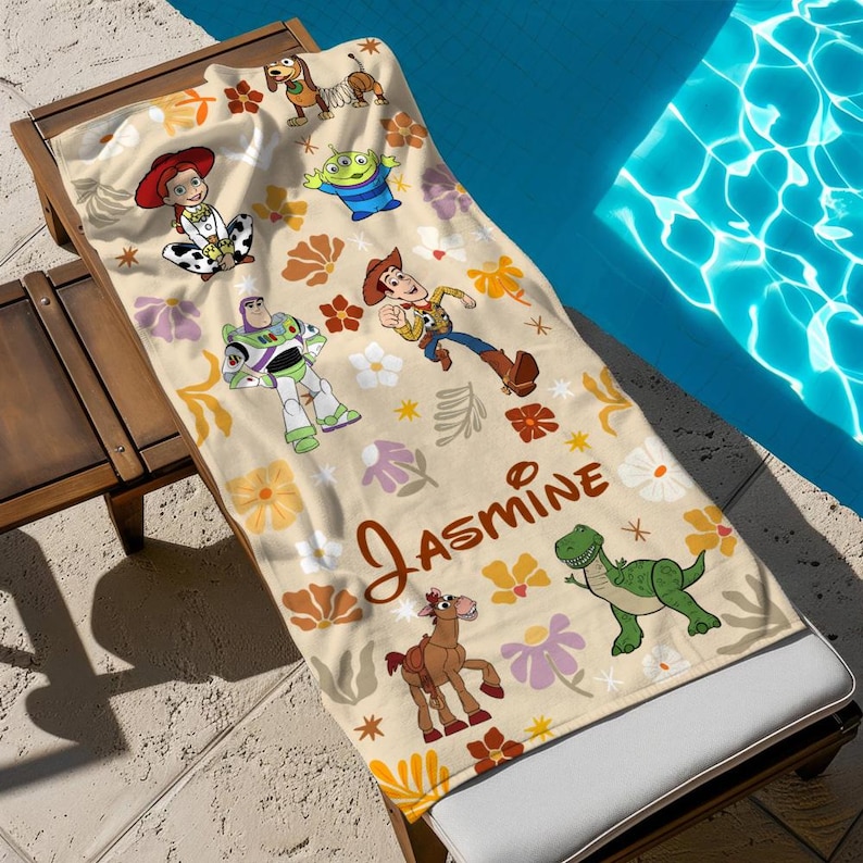 May include: A cream-colored beach towel featuring a repeating pattern of Toy Story characters including Woody, Buzz Lightyear, Jessie, Rex, Slinky Dog, and an alien, interspersed with colorful floral motifs and the name Jasmine in brown script.