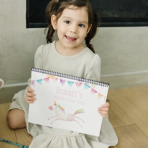 An image of a little girl holding a color book that says Scarlett's unicorn color book. Color book is a pastel unicorn under the title with a triangle banner on the top.