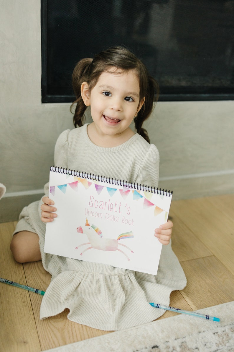 An image of a little girl holding a color book that says Scarlett's unicorn color book. Color book is a pastel unicorn under the title with a triangle banner on the top.