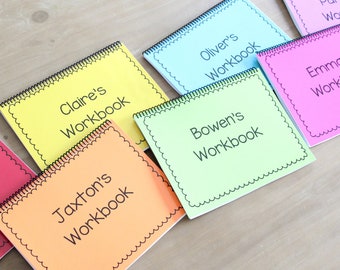 May include: A collection of colorful workbooks with black spiral binding. Each workbook has a different color cover, including yellow, orange, blue, and pink. The workbooks are labeled with names such as "Claire's Workbook" and "Jaxton's Workbook".