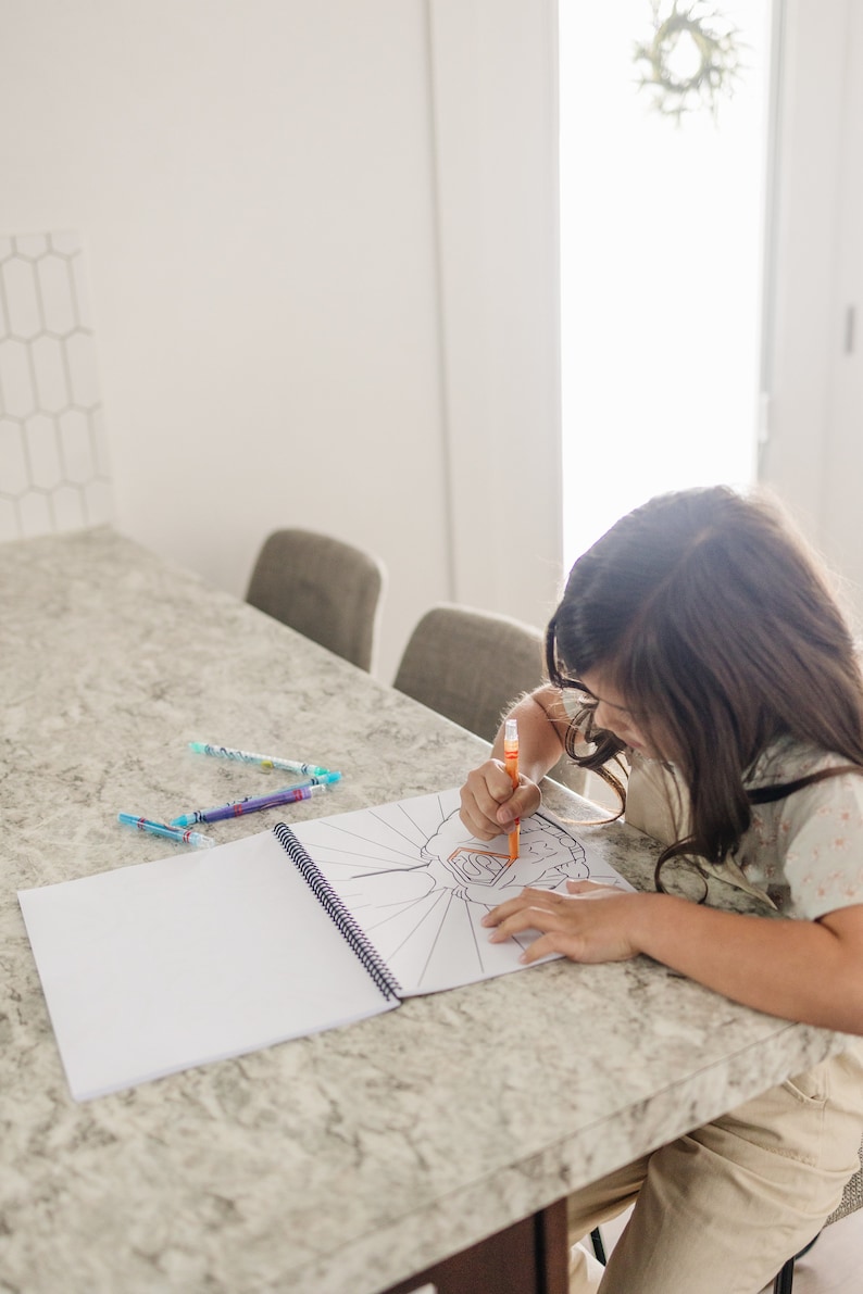 a young girl sitting at the counter coloring a super hero color book.