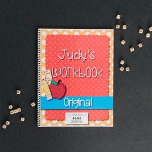 Personalized preschool workbook. The workbook says Judys workbook. It has a black background with letter dice scattered around the workbook. Workbook measures 8.5 inches by 11 inches.