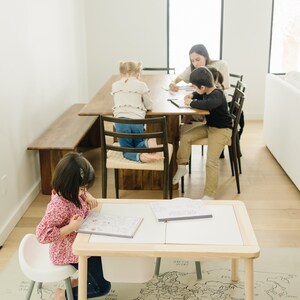 以下が含まれることがあります： 明るい木製のテーブルに、開いたノートが2冊と、世界地図のプレイマットが床に敷かれている。若い女の子がテーブルに座って、本に色を塗っている。テーブルは、大きな窓と木のベンチのある部屋にある。