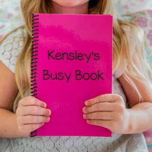 young girl holding her pink personalized laminated busy book. The book says Kensleys busy book.